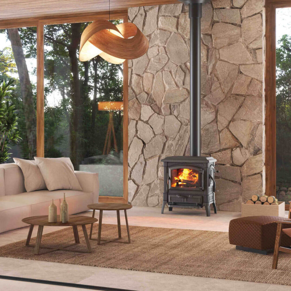 La Nordica Holzofen ISETTA CON CERCHI.16 frei stehend vor Steinwand, Feuer sichtbar, Wohnraum mit Sofa, Tisch und Fenster | Klimaworld.com