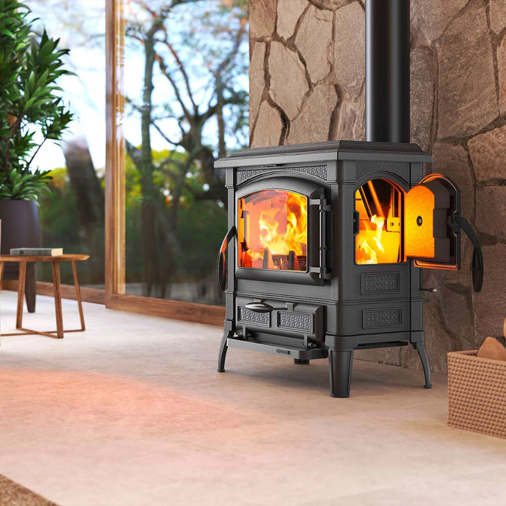 La Nordica Holzofen ISETTA CON CERCHI aus schwarzem Gusseisen mit Frontsichtfenster, offener Seitentuer, Feuer sichtbar, vor Steinwand und Fenster | Klimaworld.com