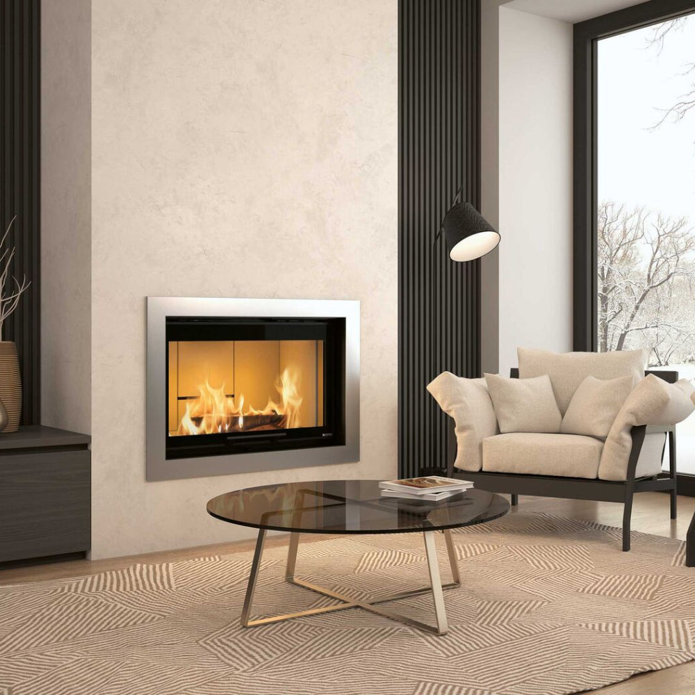 La Nordica Kamineinsatz MONOBLOCCO 90PA 2.0 in heller Wand mit brennendem Feuer, daneben Sessel und Glastisch | Klimaworld.com