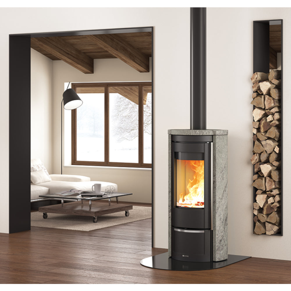Ambientebild: La Nordica Holzofen LIU' freistehend im Wohnraum, schwarzer Ofen mit Naturstein-Seiten, sichtbares Feuer, Holzregal an Wand | Klimaworld.com