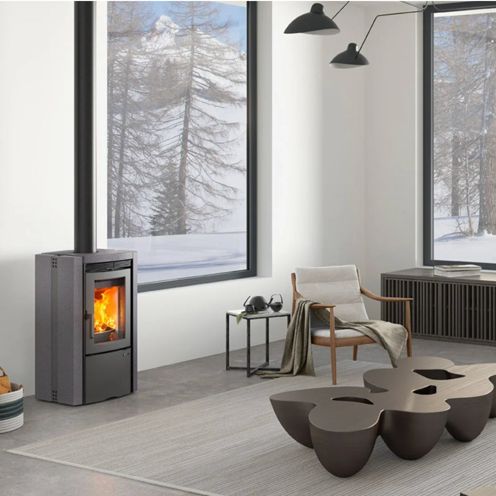 Ambientebild: La Nordica Holzofen ESTER 5.0 Vulkanstein in modernem Wohnzimmer, grauer Ofen mit sichtbarer Flamme am Fenster | Klimaworld.com