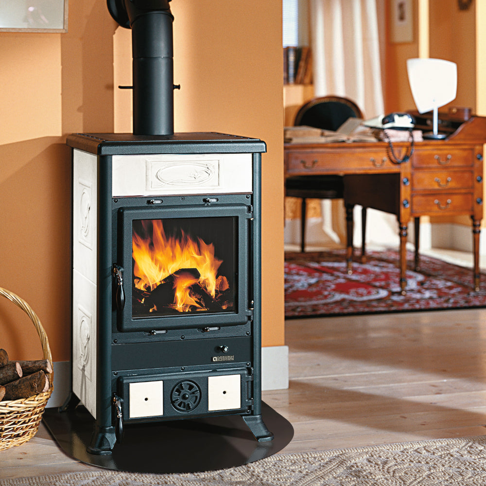 Ambientebild: La Nordica Holzofen ROSSELLA R1.16 CERAMICA, Korpus schwarz, verzierte weisse Keramikseiten, Glasfenster vorn mit Feuer vor beiger Wand | Klimaworld.com