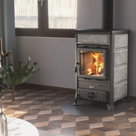 Ambientebild: La Nordica Holzofen ROSSELLA R1.16 Petra Kaminofen, grauer Naturstein, Glasfenster mit Feuer, im Raum mit Fenster, Bodenmuster | Klimaworld.com