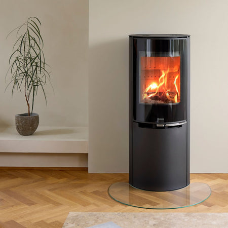 Ambientebild: Aduro Kaminofen 9.8 Lux, schwarzer runder Ofen mit Glasfront, brennendes Holz im Wohnraum neben Pflanze | Klimaworld.com