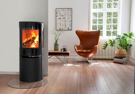 Ambientebild: Aduro Hybridofen H3 Lux in modernem Wohnraum auf Glasplatte, daneben Ledersessel, Tisch und Pflanze am Fenster | Klimaworld.com