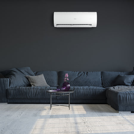Ambientebild: Haier Klimaanlage Wandgeraet Pearl Premium ueber blauem Ecksofa an dunkler Wand, Tisch mit Vase davor | Klimaworld.com
