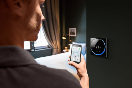 Ambientebild: Daikin Madoka Fernbedienung an Schlafzimmerwand hängend mit Bediener am Smartphone | Klimaworld.com