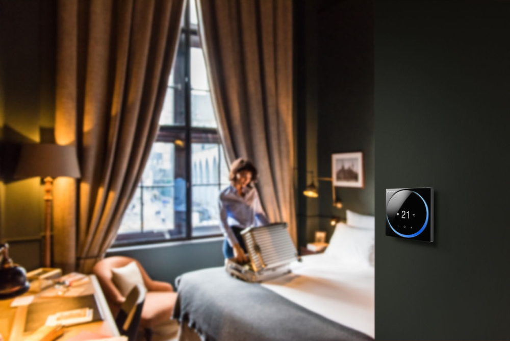 Ambientebild: Daikin Madoka Fernbedienung an Wand hängend im Hotelzimmer, Frau mit Koffer | Klimaworld.com