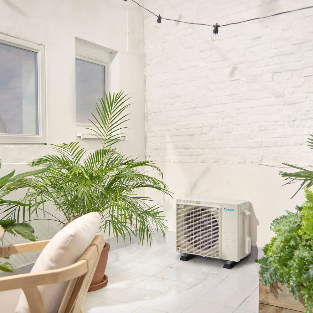 Ambientebild: Daikin Sensira Aussengeraet auf heller Terrasse vor weisser Ziegelwand, umgeben von Palmen und Pflanzen | Klimaworld.com
