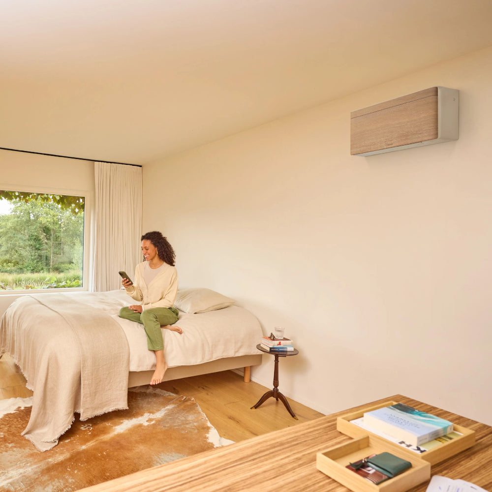 Ambientebild: Daikin Stylish FTXA-DP Light Wood Wandgeraet ueber einem Bett, Frau mit Smartphone und Fensterblick | Klimaworld.com
