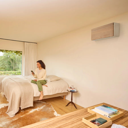 Ambientebild: Daikin Stylish FTXA-DP Light Wood Wandgeraet ueber einem Bett, Frau mit Smartphone und Fensterblick | Klimaworld.com
