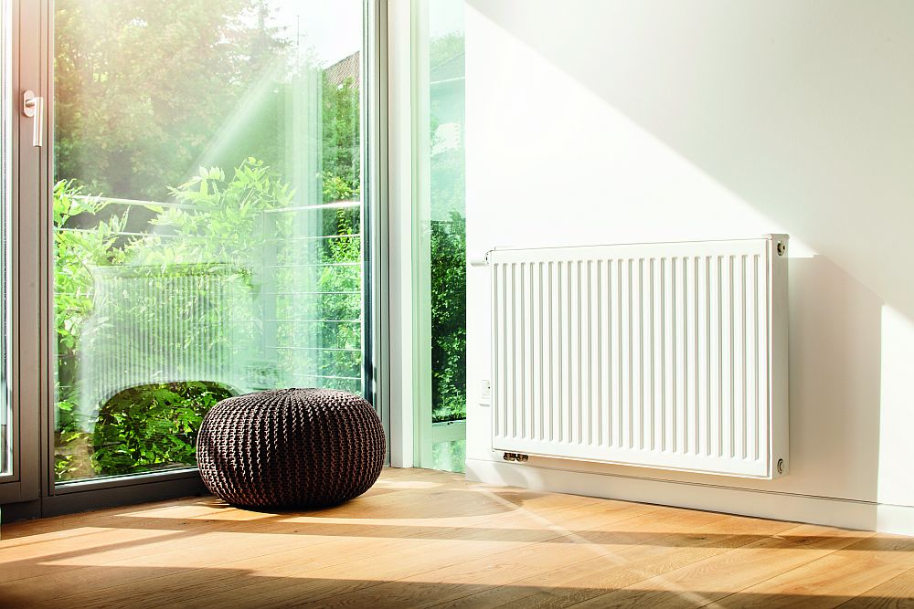 Weisser Flachheizkoerper an der Wand in einem lichtdurchflutetem Raum, neben einem Pouf, Ventil links | klimaworld.com