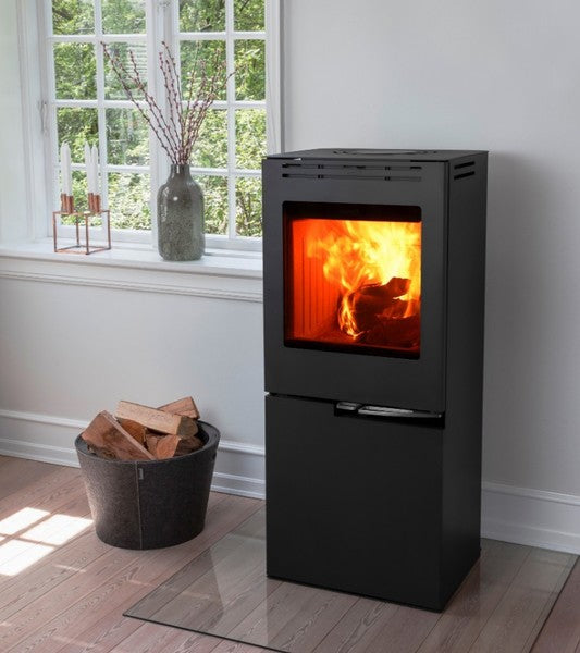 Ambientebild: Schwarzer Aduro Hybridofen H5 auf Glasplatte, mit brennendem Feuer im Wohnraum neben Fenster und Holzkorb | Klimaworld.com